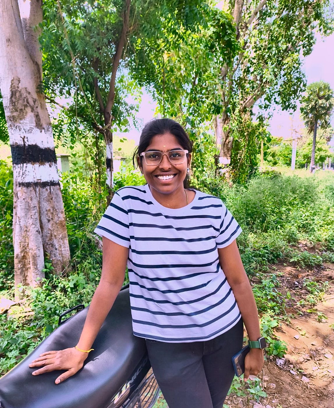 Kaviya in glasses and a striped tee, smiling under trees